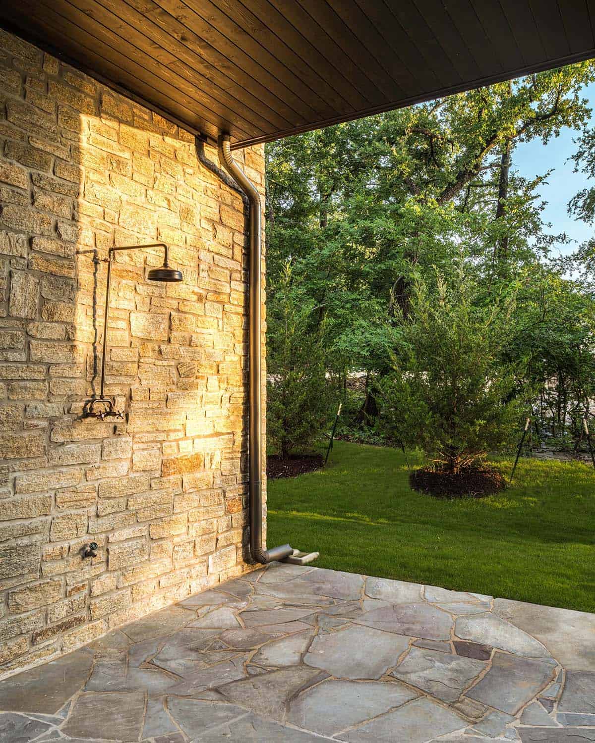 Outdoor stone shower fixture mounted on a stone wall under a covered porch, overlooking a grassy yard
