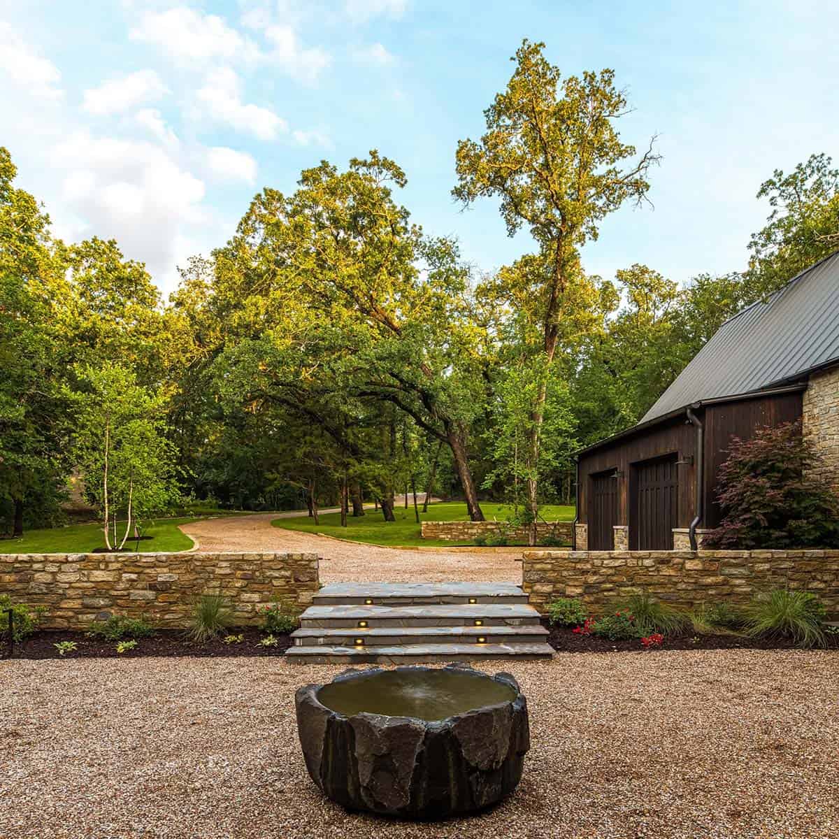 Gravel driveway curving through trees toward a dark wood garage, with a carved stone water basin and stone steps in the foreground