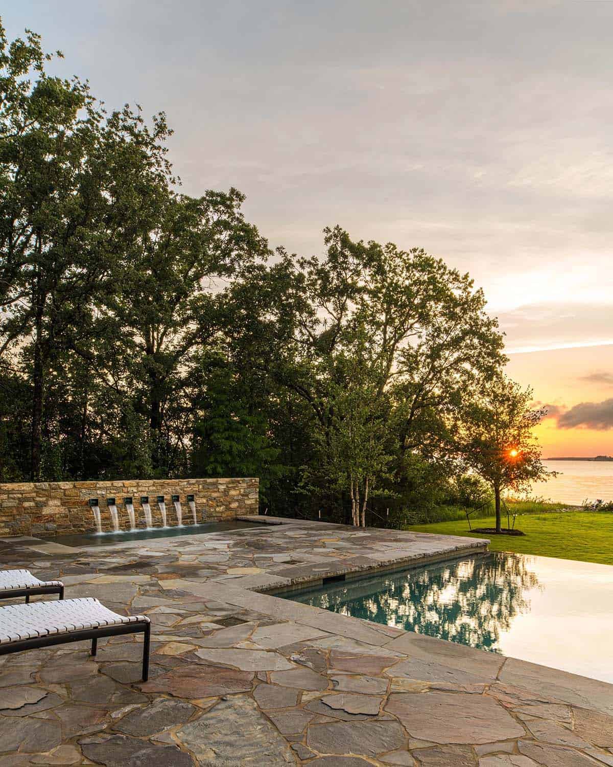 Infinity-edge pool with a stone spillway wall, lounge chairs, and a lake sunset in the background