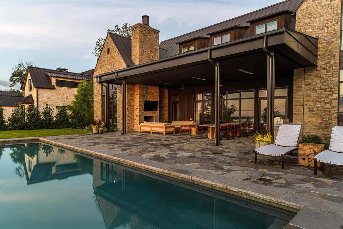 Covered outdoor living area with stone fireplace, TV, and dining table beside a long pool reflecting the house