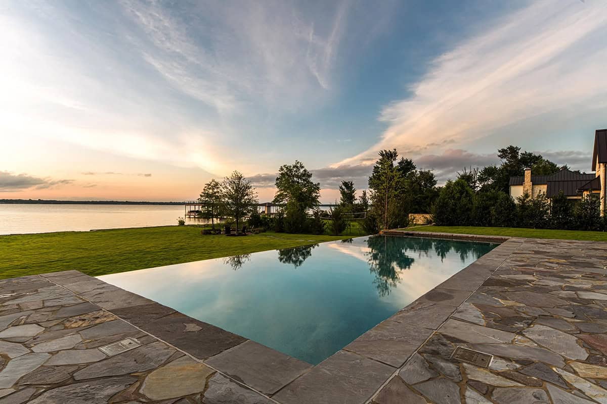 Infinity-edge pool overlooking a grassy lawn and lake at sunset, framed by a stone patio