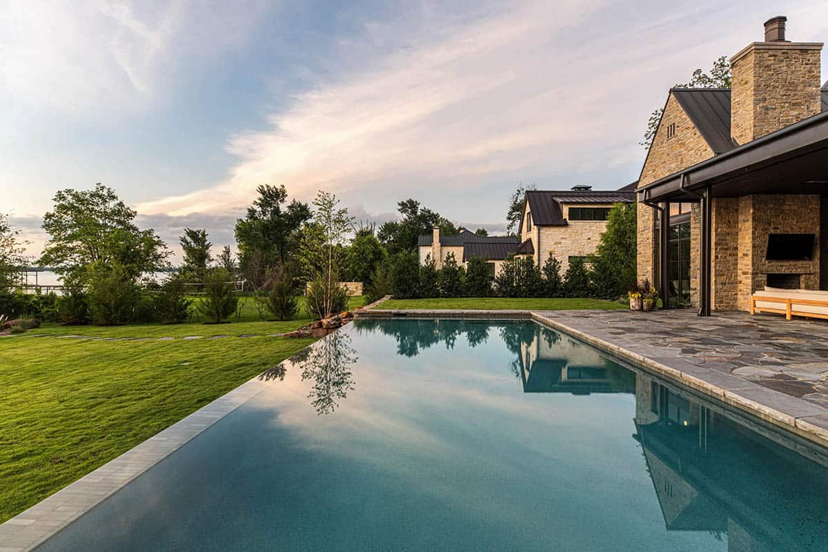 Pool deck view of the stone lake house with chimney, covered porch, and lawn leading to the water