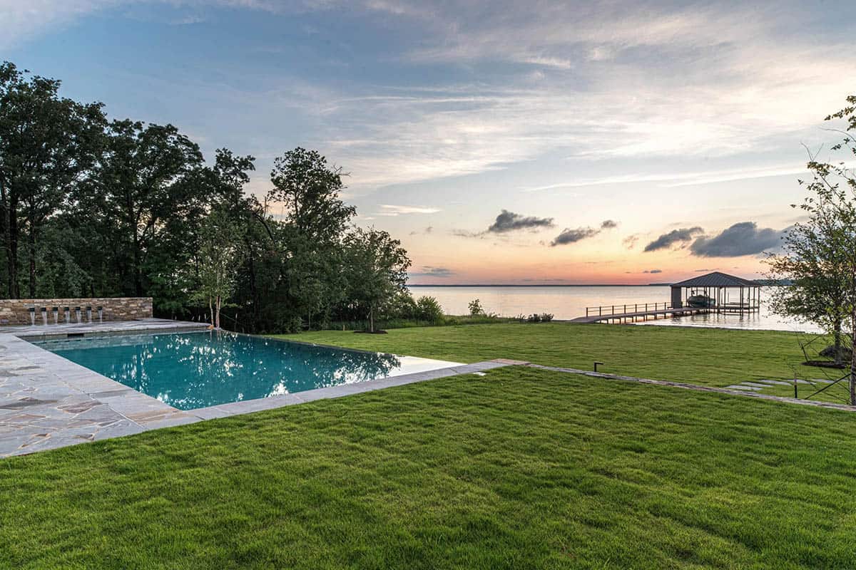 Infinity pool beside a manicured lawn leading to a lakeside dock at dusk
