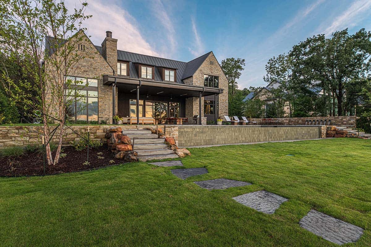Rear elevation of the stone and metal-roofed house with covered porch, stone retaining walls, and a green lawn with stepping-stone path