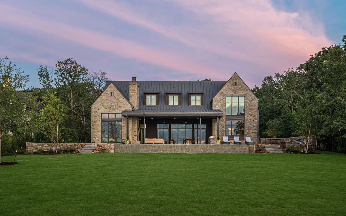 Twilight view of the rear facade with pink sky, stone gables, covered porch, and broad lawn