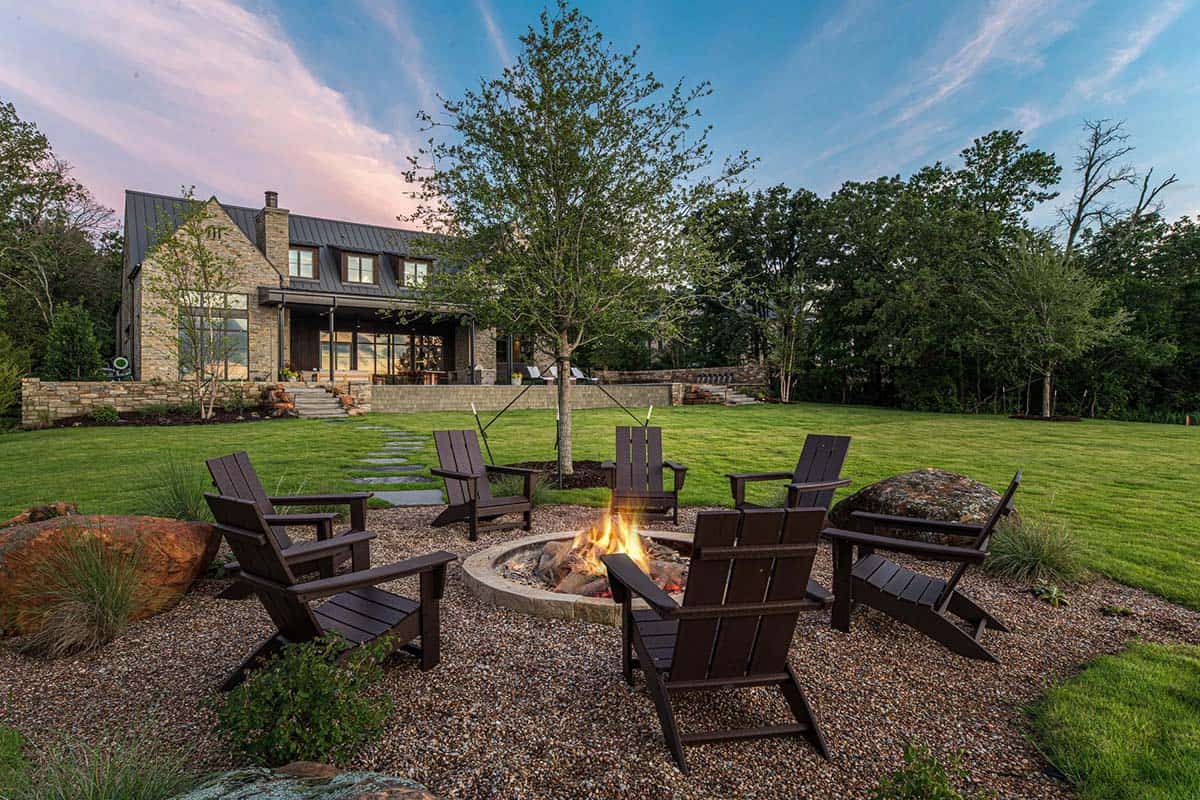 Fire pit with dark Adirondack chairs on a gravel patio facing the lake house lawn at twilight
