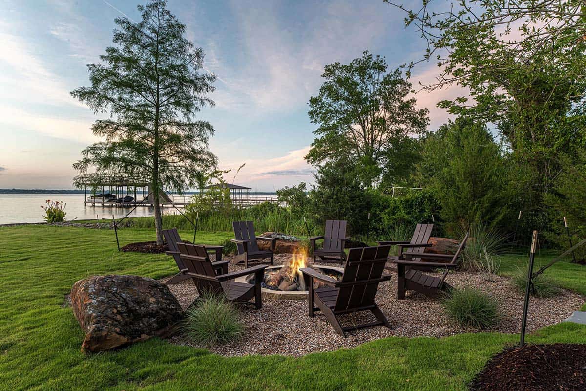 Adirondack chairs around a lit fire pit overlooking a lake with a boat dock at sunset