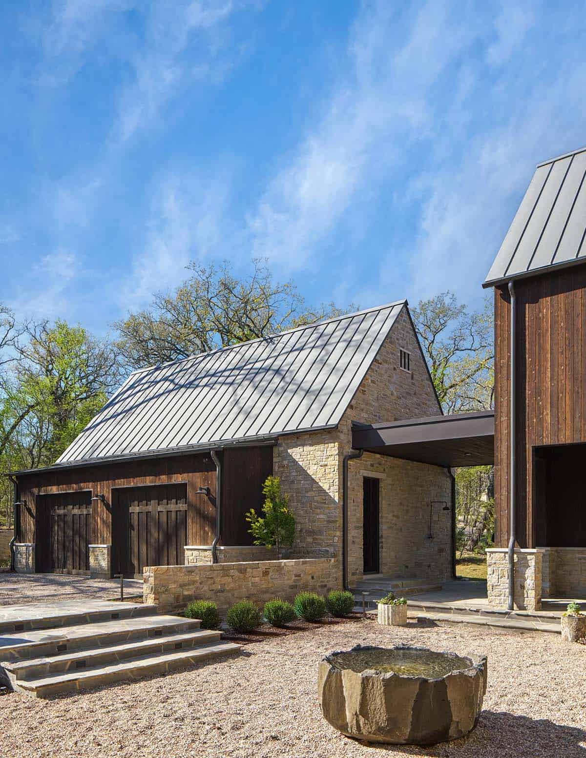 Garage and outbuilding with board-formed dark wood siding, stone base, metal roof, and a carved stone water basin in the gravel courtyard