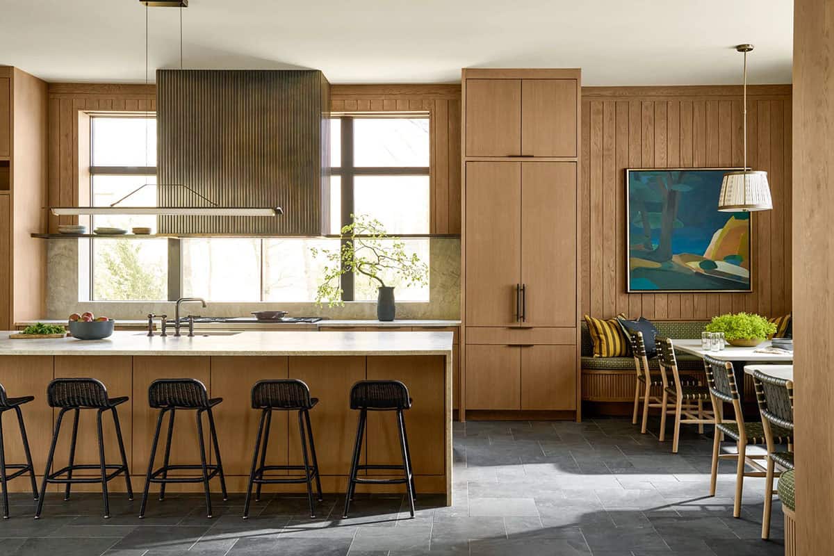 Open kitchen with wood cabinetry, a fluted brass-toned range hood, black bar stools, and a banquette dining nook