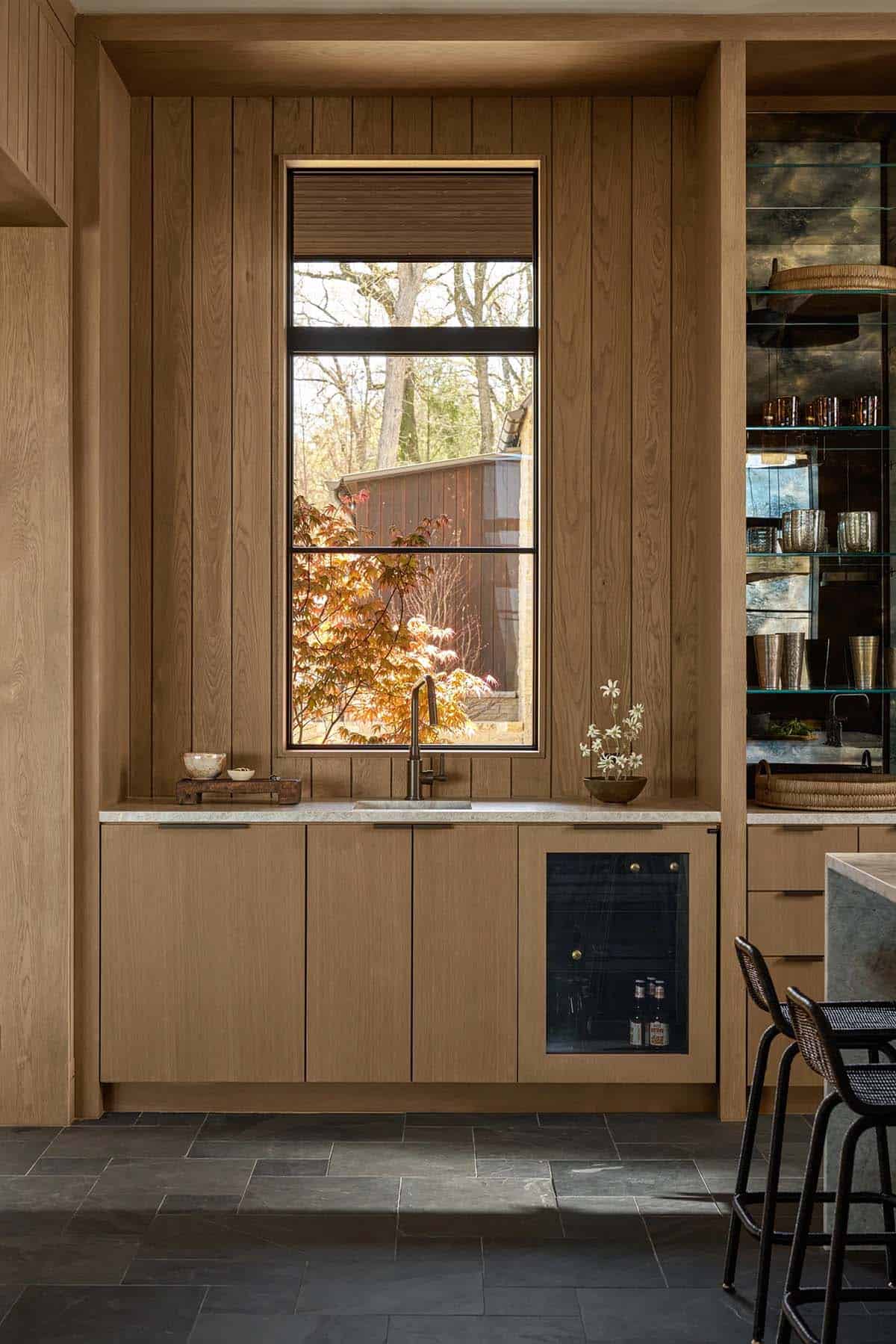 Wood-paneled wet bar with a sink, wine fridge, and mirrored glass shelving beside a window framing a Japanese maple