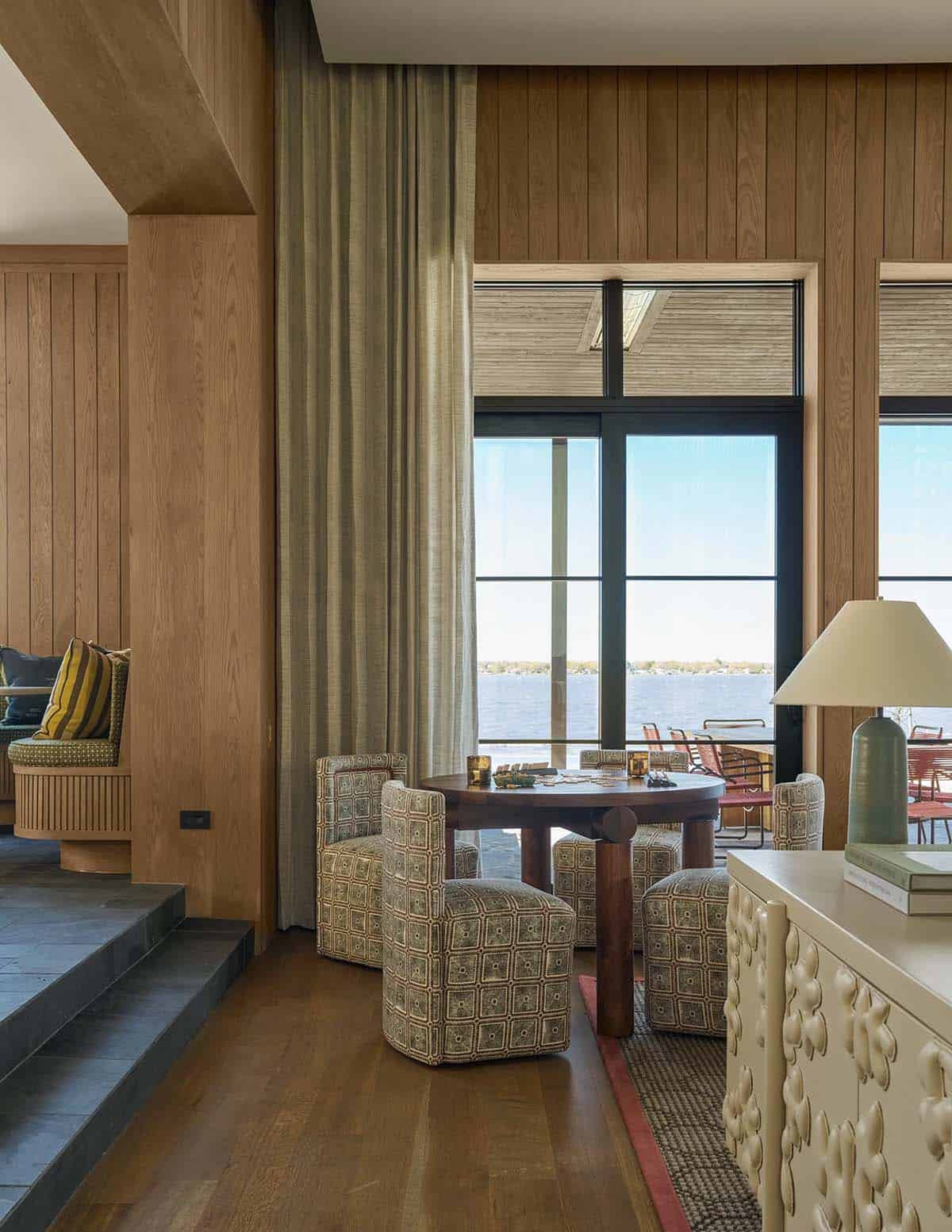 Wood-paneled sitting nook with a round game table, patterned chairs, and floor-to-ceiling windows overlooking the lake