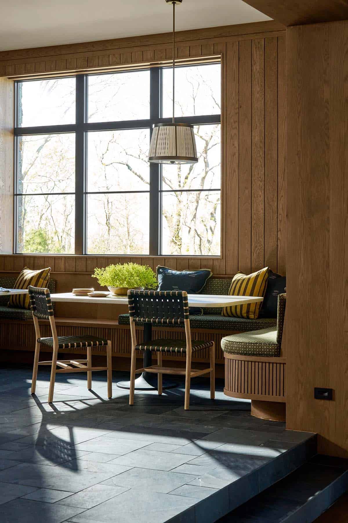 Close-up of the dining banquette with green cushions, woven chairs, and a pendant light against large windows