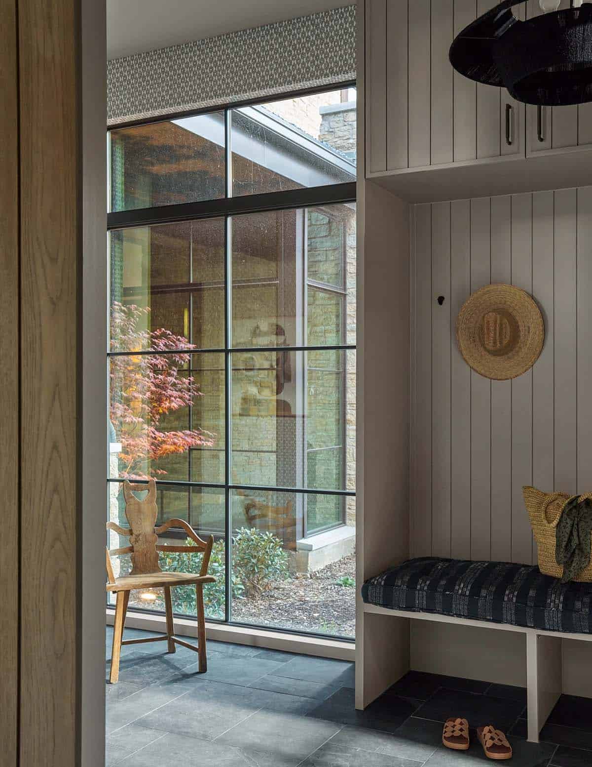 Mudroom nook with built-in bench, woven bag, and straw hat, beside a glass hallway view of a courtyard Japanese maple