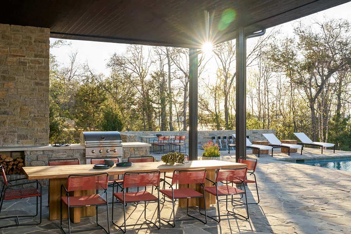 Covered outdoor kitchen and dining area with a built-in grill, wood table with red chairs, and pool loungers beyond