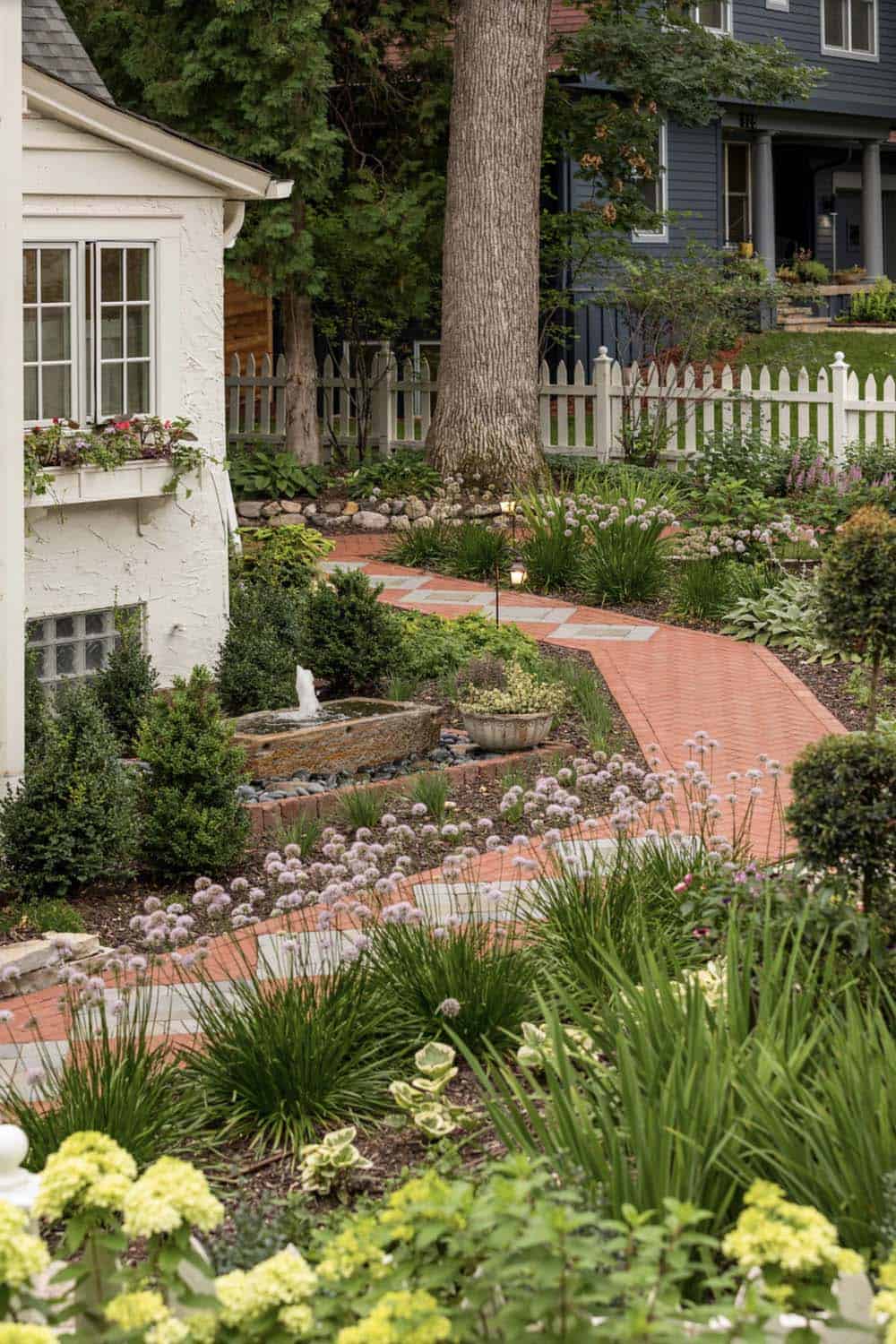 Curved brick walkway through layered cottage garden with stone fountain and white picket fence