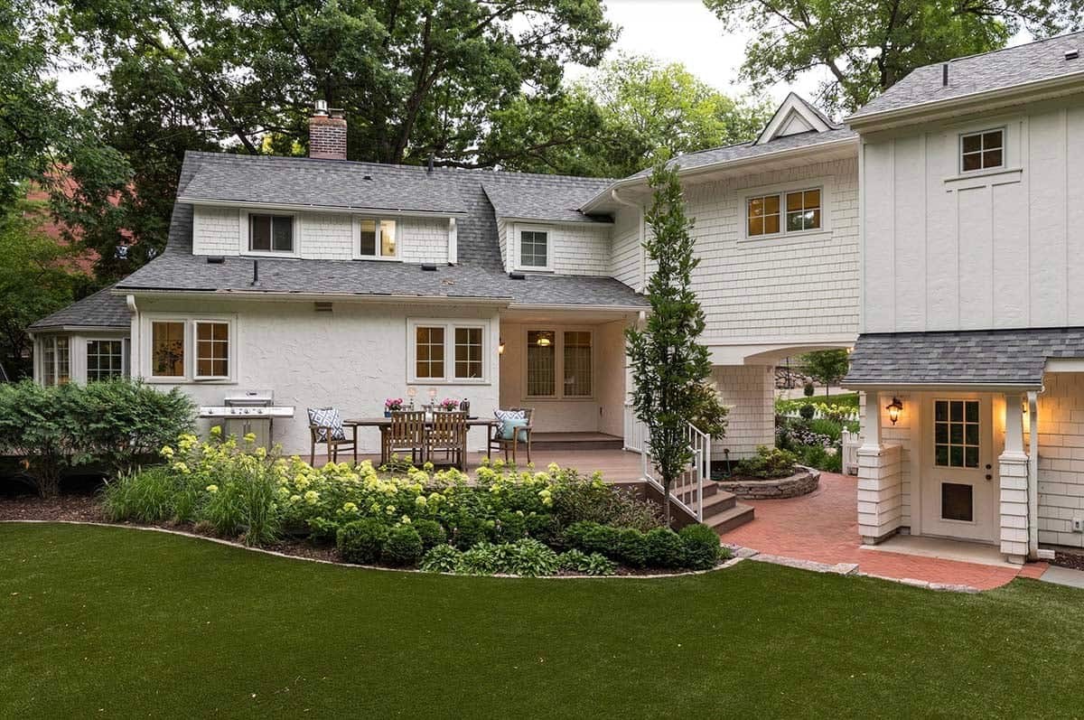 Rear view of white cottage home with deck dining area, hydrangea beds, and brick courtyard