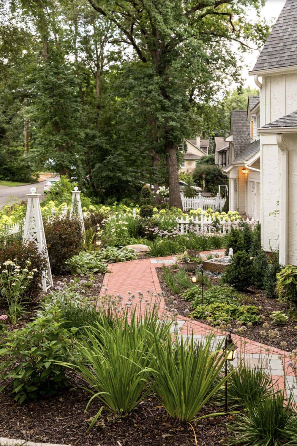 Brick garden path through layered perennial beds with white obelisk trellis and path lighting