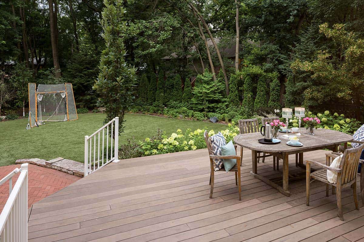 Backyard deck with round teak dining table overlooking lawn, lacrosse net, and hydrangea garden beds