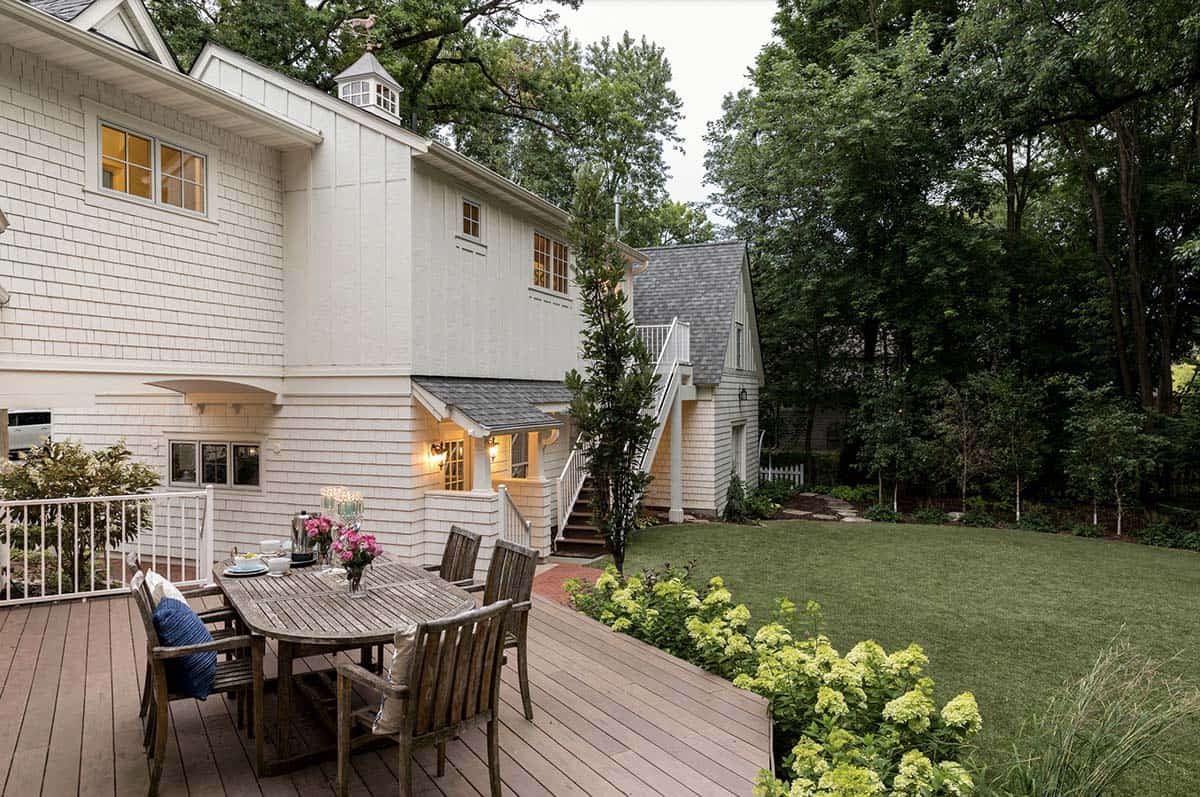Rear view of white shingle cottage home with deck dining set, carriage house, and hydrangea border at dusk