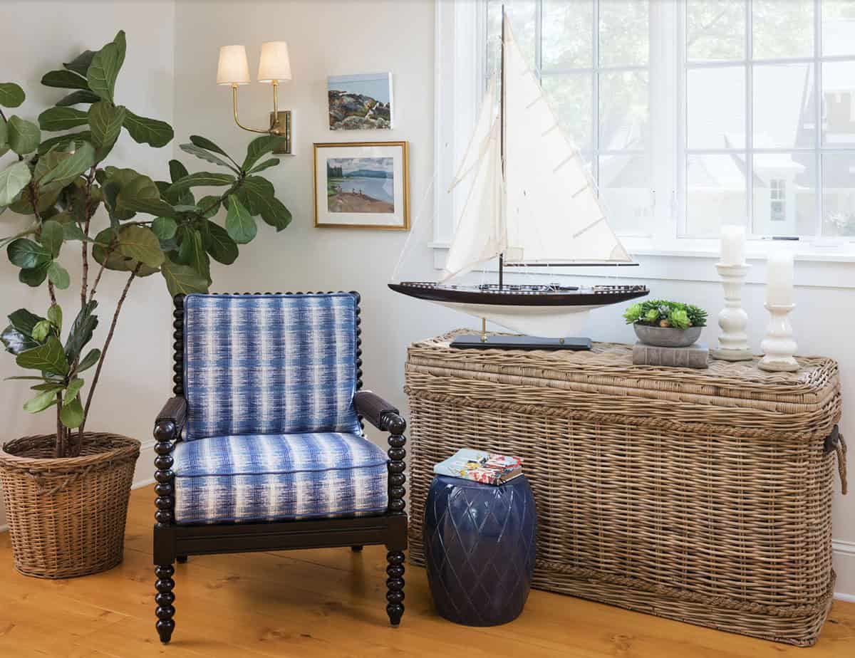 Blue ikat spool chair beside wicker trunk with model sailboat, fiddle leaf fig, and coastal artwork