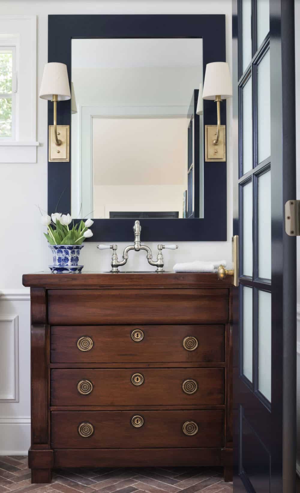 Powder room with antique wood vanity, navy mirror surround, brass sconces, and bridge faucet