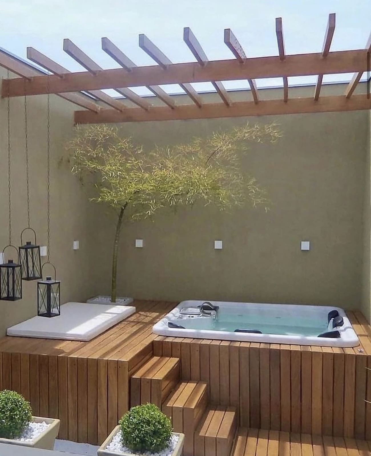 A raised wood deck hot tub with a closed cover platform, pergola, hanging lanterns, and an indoor olive tree against a courtyard wall