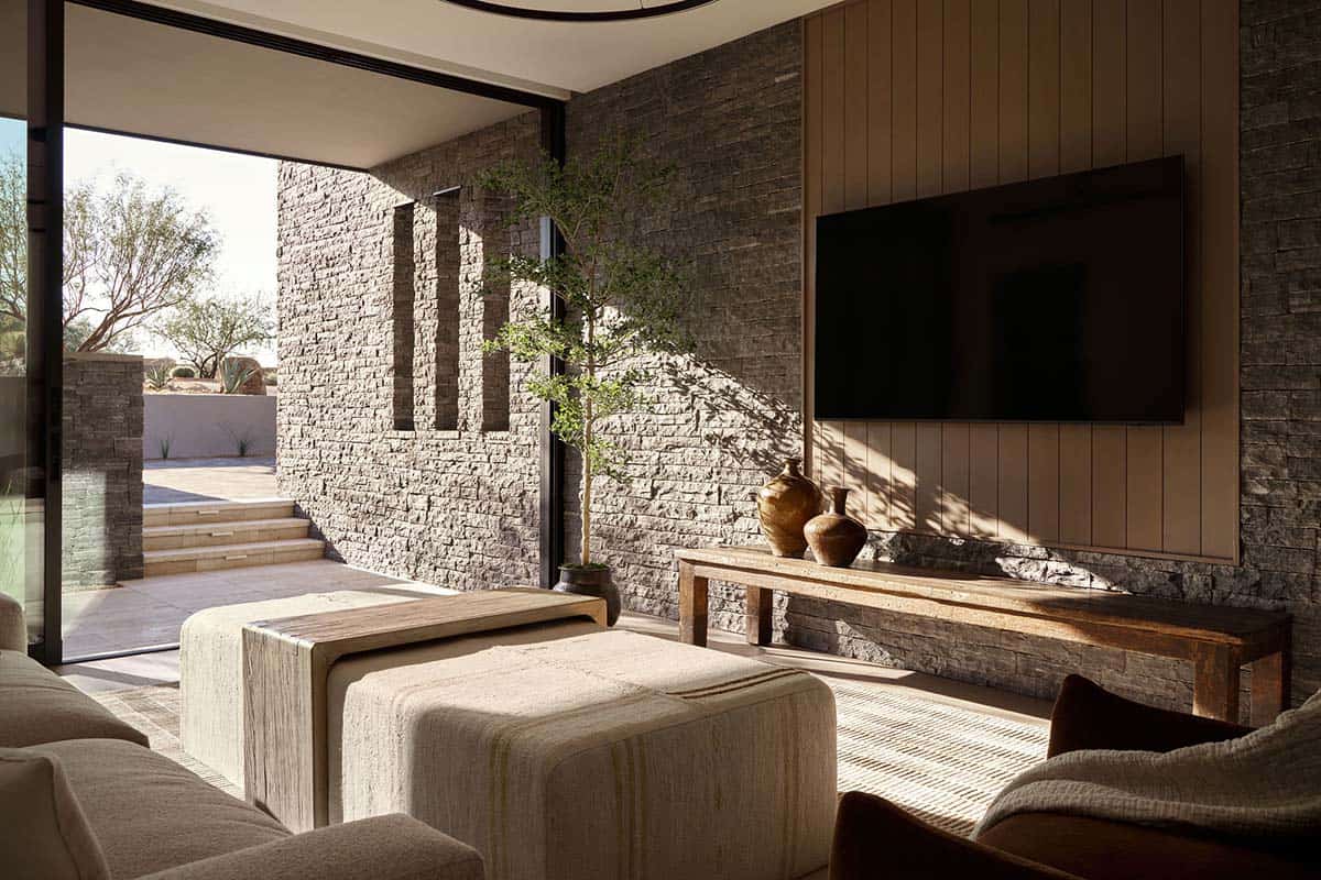 Living room with stacked stone walls, wood-panel TV surround, travertine console, and sliding glass doors opening to a desert courtyard