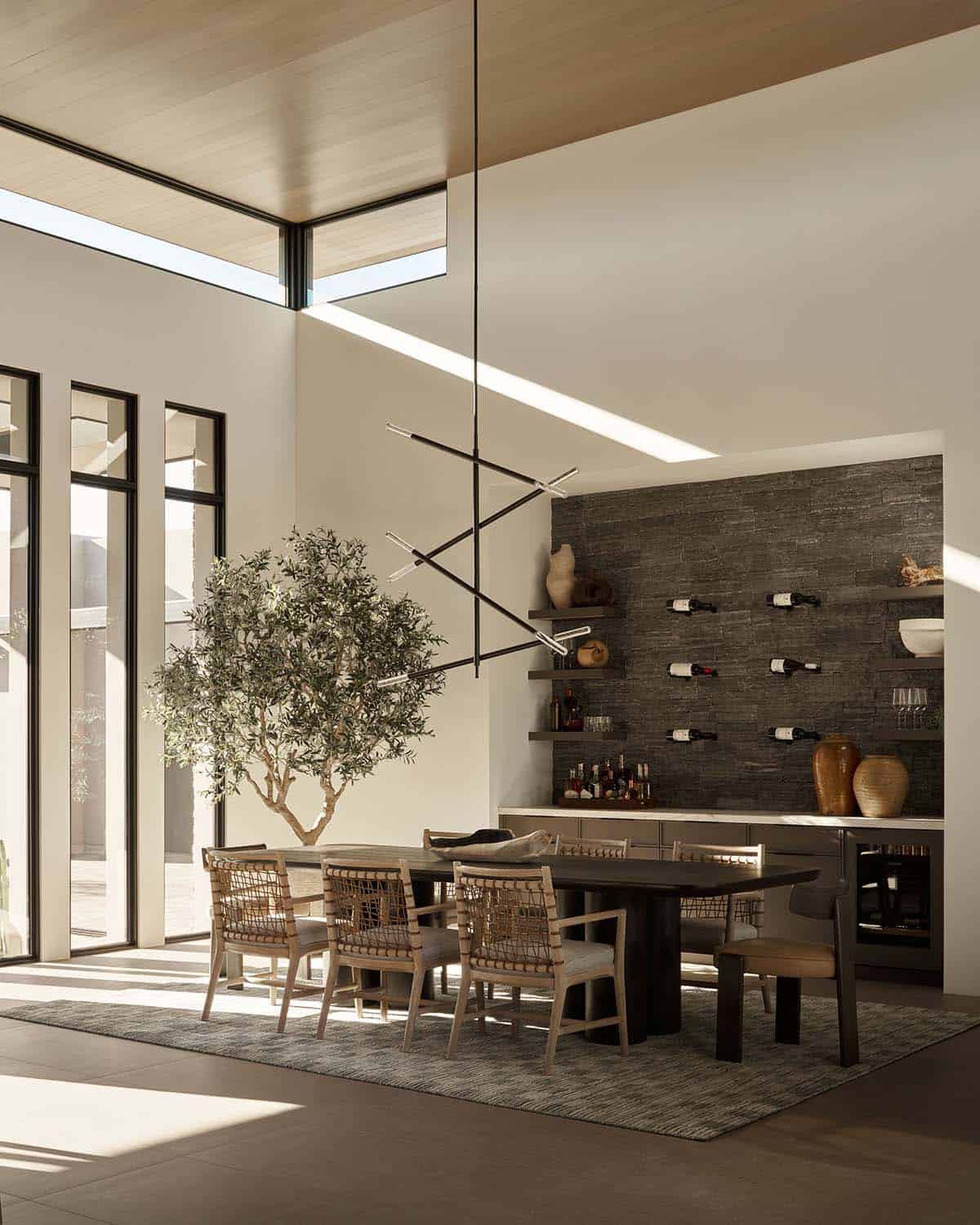 Dining room with double-height ceilings, sculptural linear chandelier, dark wood table, woven chairs, olive tree, and built-in wine wall on stacked stone