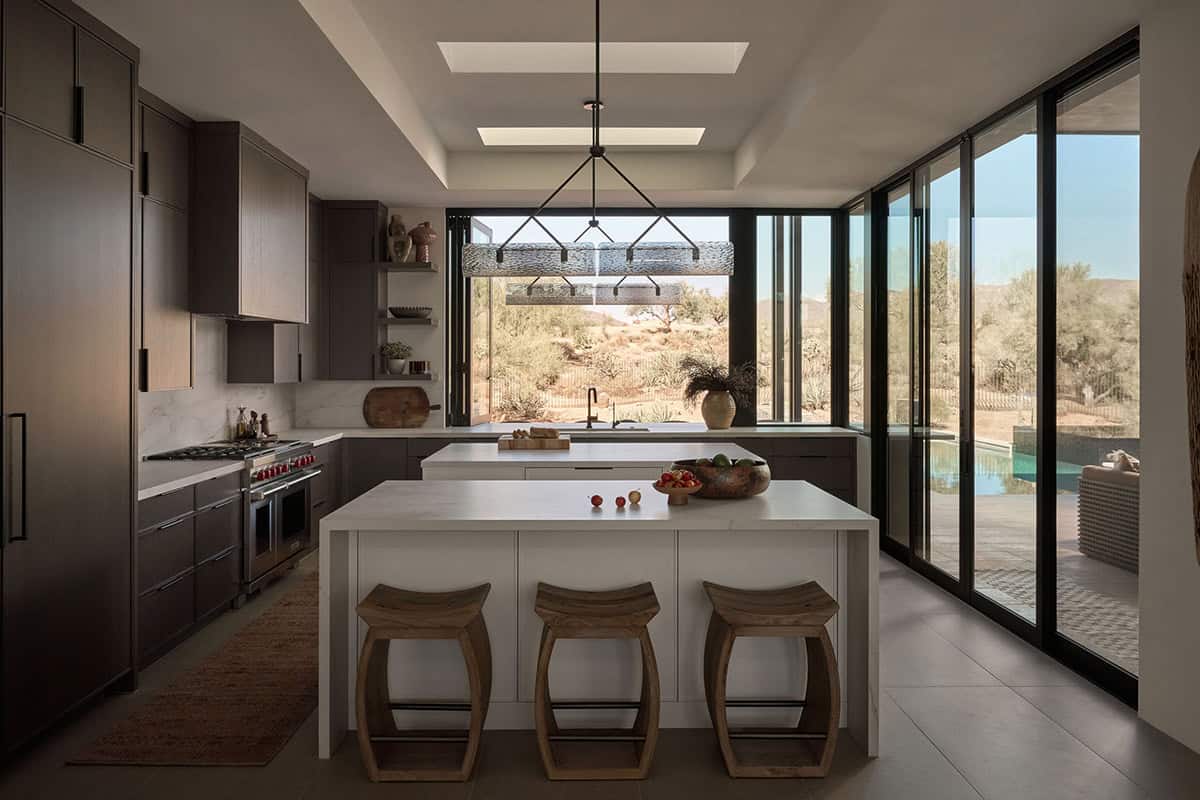 Desert modern kitchen with dark wood cabinetry, white quartz island, bar stools, pro range, skylights, and sliding glass doors with Sonoran Desert views