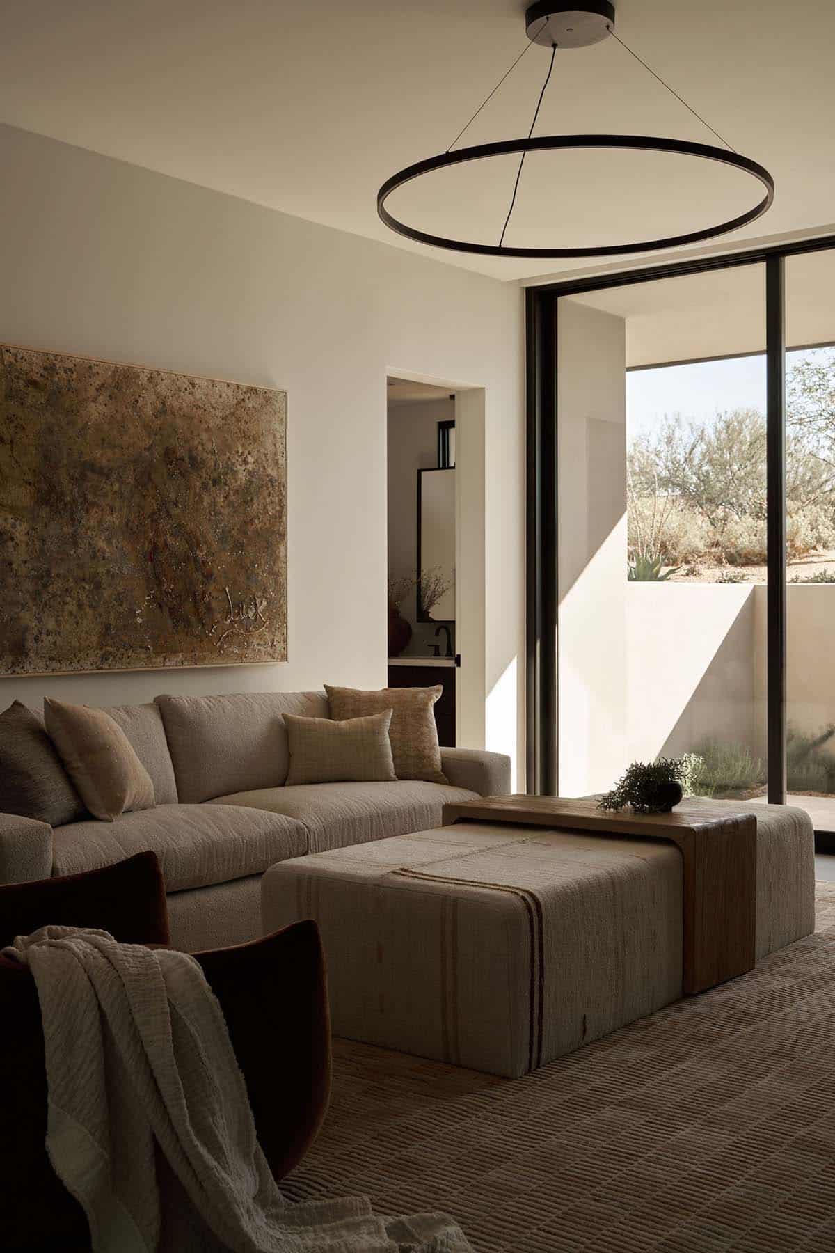 Sitting room with neutral sectional, linen ottoman, abstract canvas, circular pendant light, and sliding glass door to a walled desert courtyard