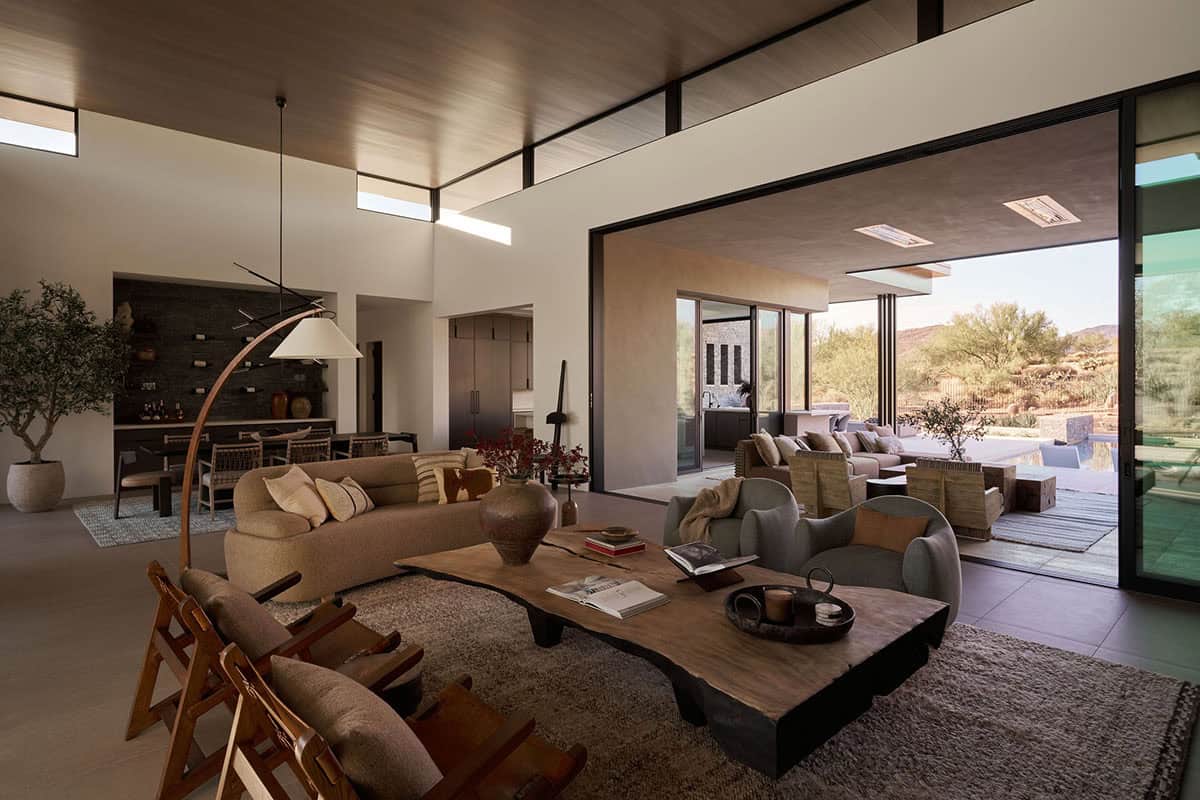 Open-plan living and dining room with wood ceiling, live-edge coffee table, and panoramic sliding doors opening to a covered desert patio