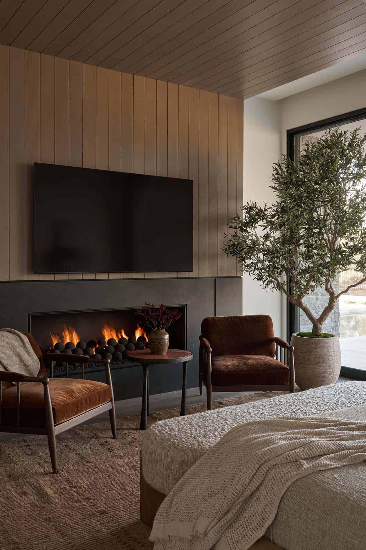 Primary bedroom sitting area with wood-slat ceiling, linear fireplace, velvet armchairs, olive tree, and sliding glass door to a walled exterior