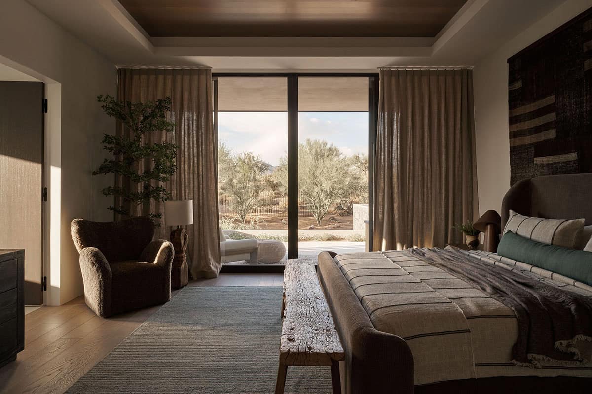 Guest bedroom with tray ceiling, linen drapery, shearling chair, raw wood bench, and floor-to-ceiling sliding doors framing desert landscape views
