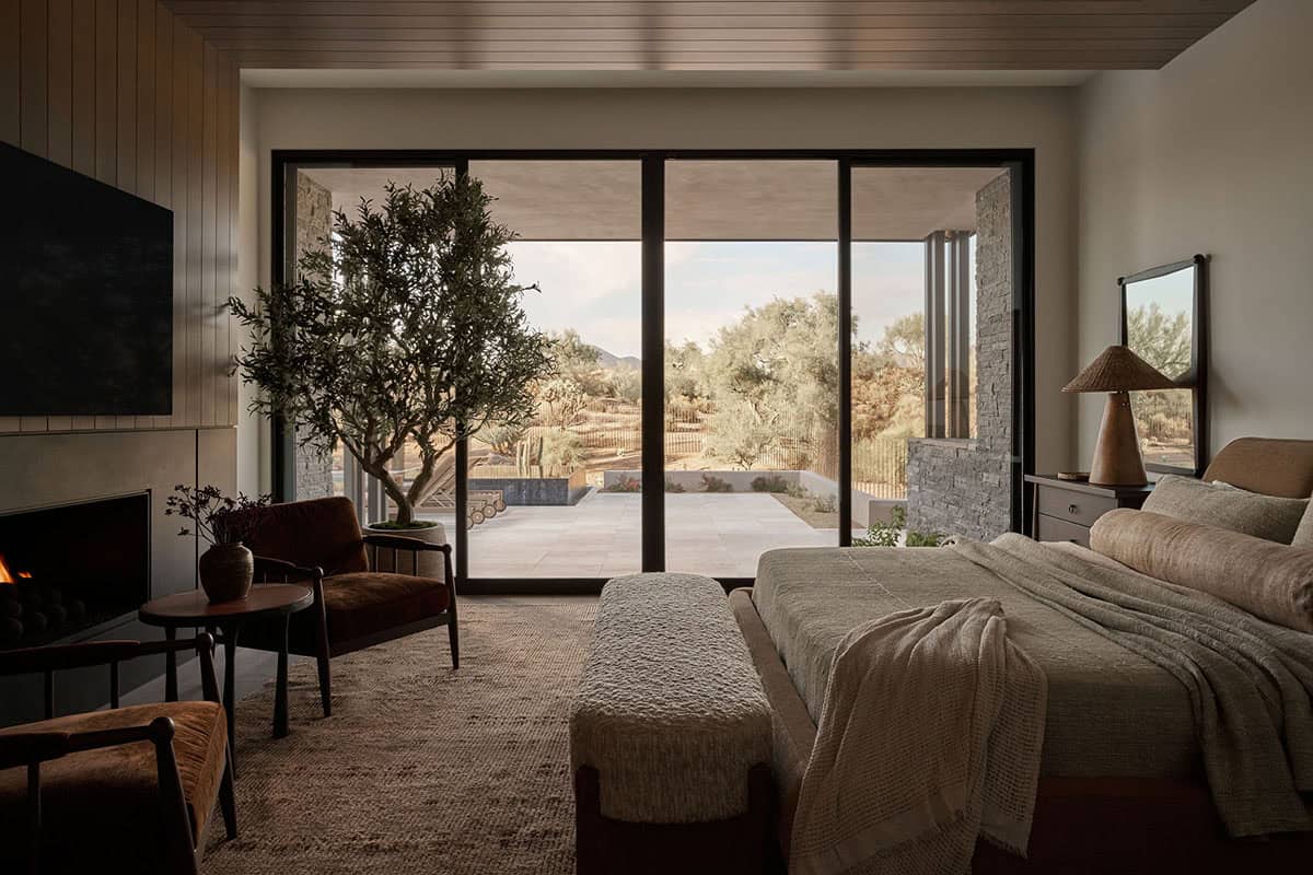 Primary bedroom with wood-slat ceiling, linear fireplace, velvet chairs, upholstered bench, and wide sliding glass doors opening to a stone patio
