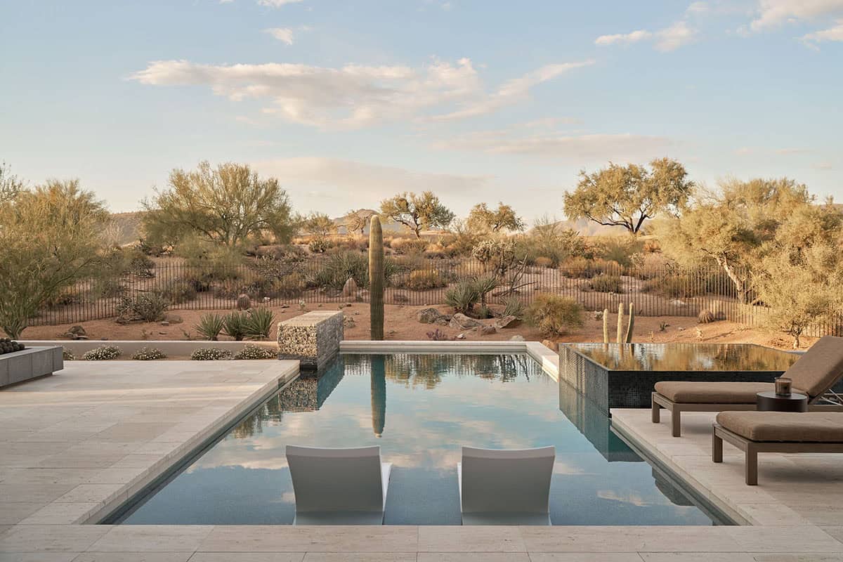 Rectangular pool with in-water lounge chairs, gabion water feature, stone coping, and open Sonoran Desert views at golden hour
