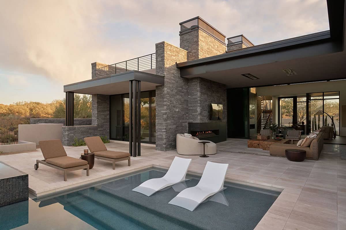 Pool and covered patio at dusk with stacked stone exterior, linear outdoor fireplace, in-pool sun loungers, and lit interior visible through glass walls