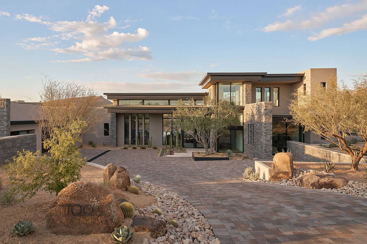 Front exterior of desert modern home with stacked stone columns, flat rooflines, floor-to-ceiling glass, and native desert landscaping