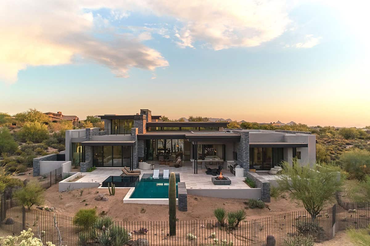 Aerial view of desert modern home at sunset with flat roof, stacked stone accents, rectangular pool, fire pit, and Sonoran Desert surroundings