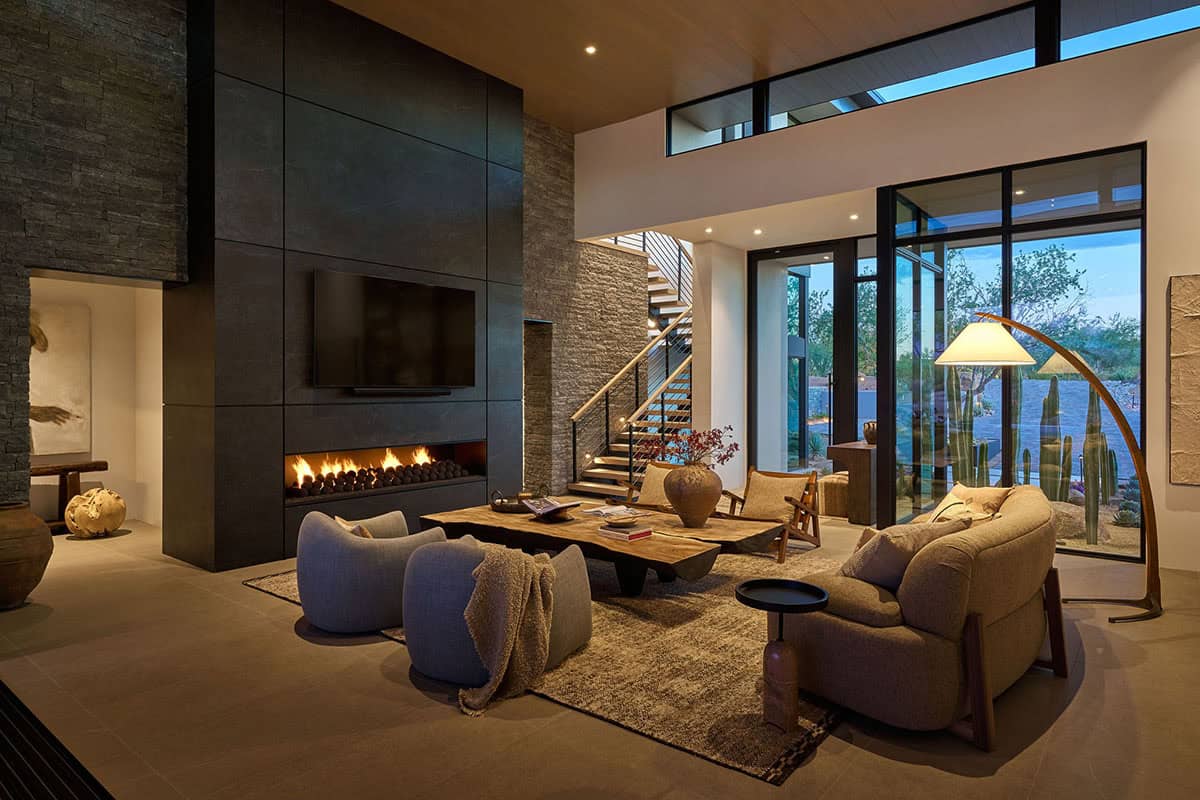 Great room at night with floor-to-ceiling stone fireplace wall, linear gas insert, live-edge coffee table, open staircase, and floor-to-ceiling glass overlooking the desert