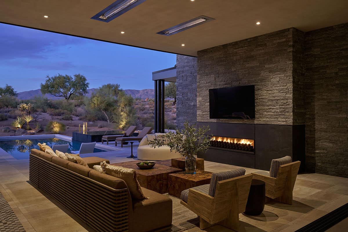 Covered outdoor living room at night with stacked stone fireplace wall, linear gas insert, linen sectional, wood coffee tables, pool, and mountain views