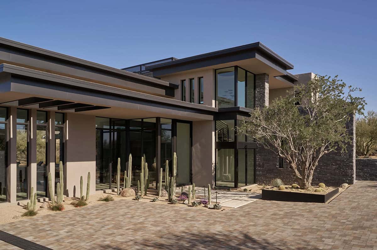 Desert modern home exterior with flat rooflines, steel-framed floor-to-ceiling glass, stacked stone accents, columnar cacti, and a desert tree courtyard under a clear blue sky