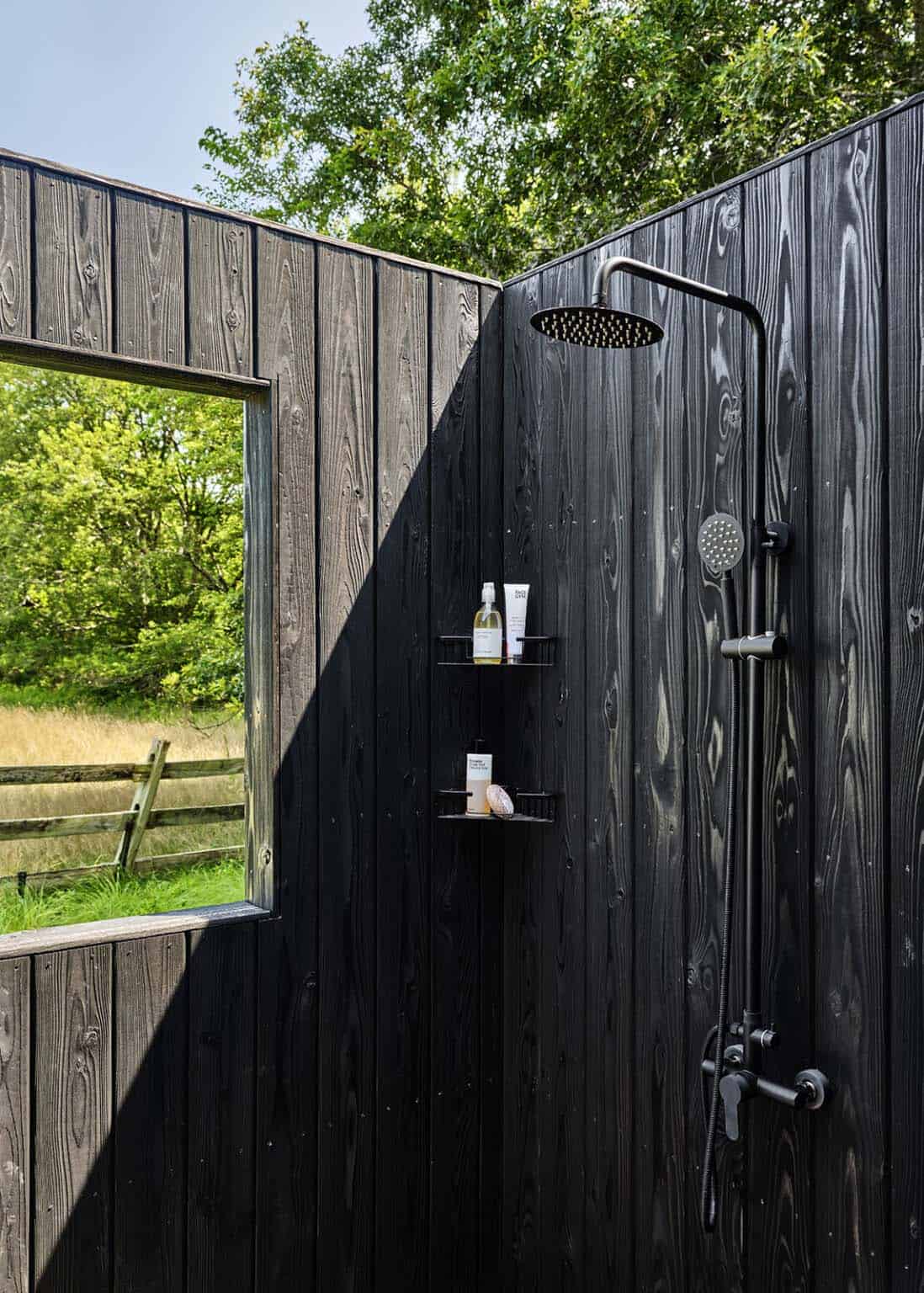 Outdoor shower with black wall-mounted fixtures against charred wood siding and a meadow view