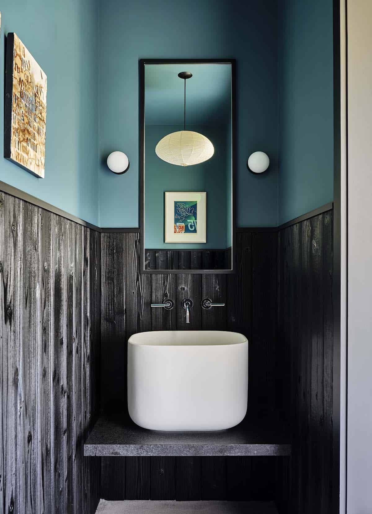 Small powder room with teal walls, black wood paneling, oval white vessel sink, and round mirror