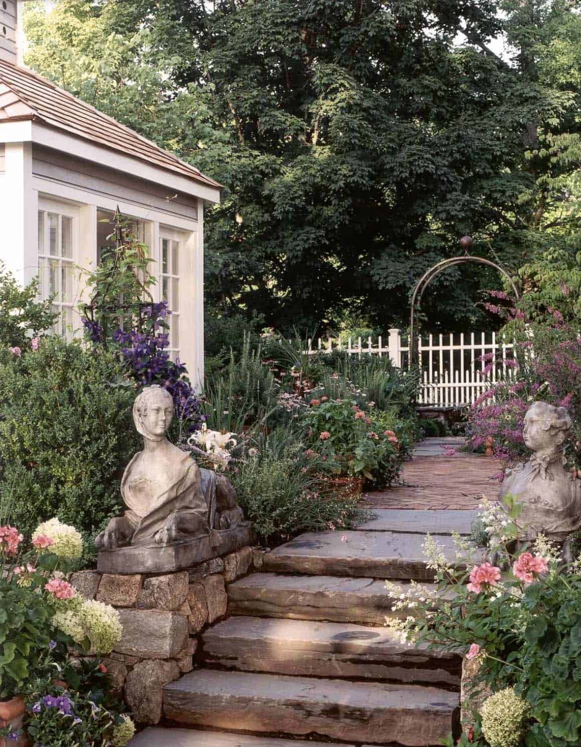 Stone steps and flagstone path lined with classical statues, layered cottage garden borders, and a metal rose arch in a small English backyard
