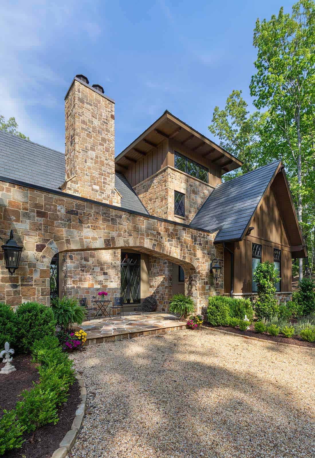 Stone turret and arched entry of European-style lake house with gravel driveway and lush landscaping