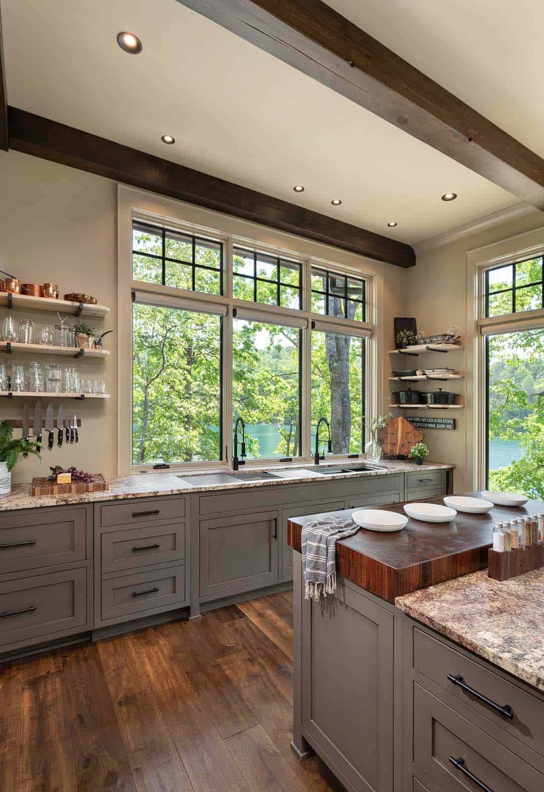 Kitchen sink wall with sage green cabinets, granite counters, exposed wood beams, and large lake-view windows