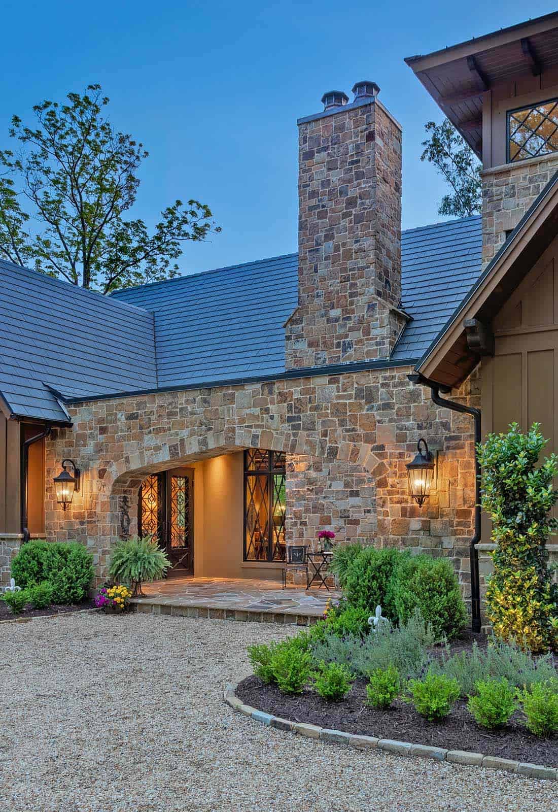 Stone entry arch at dusk with lantern sconces, flagstone patio, and manicured shrub landscaping