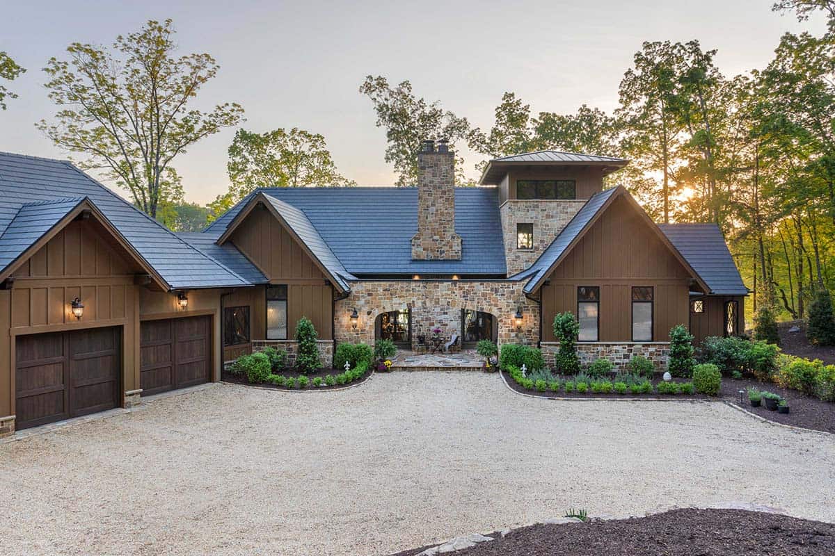 Full front elevation of European lake house at sunset with double garage, stone chimney, and metal roof
