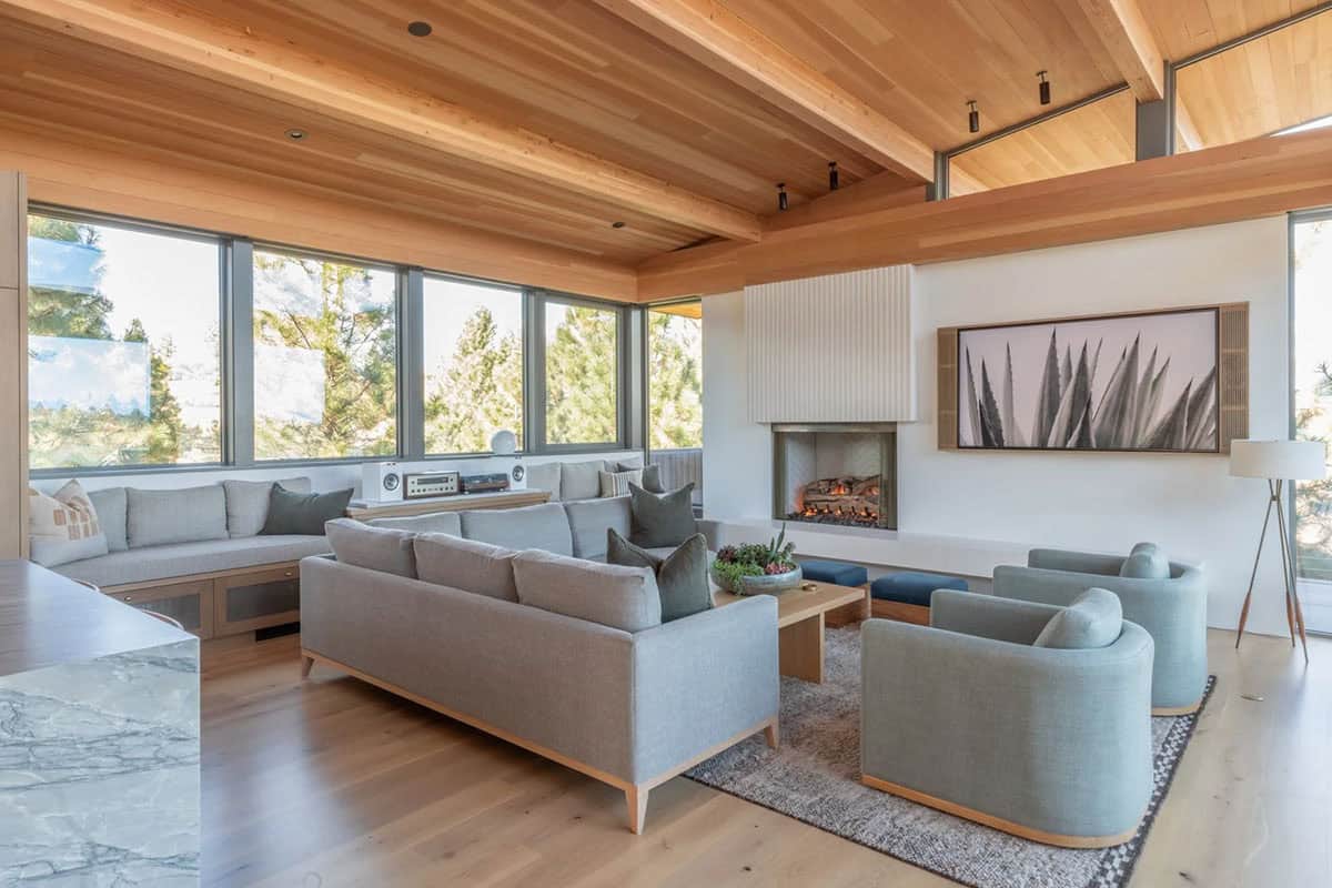 Living room with wood beam ceiling, fireplace, and wraparound windows framing ponderosa pines