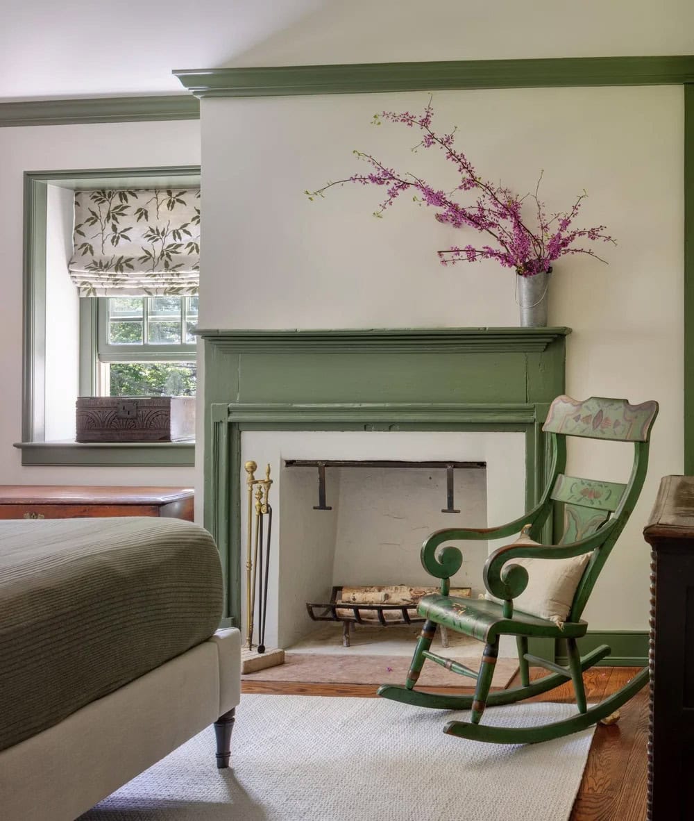 Bedroom corner with sage-green fireplace mantel, painted rocking chair, and flowering branch
